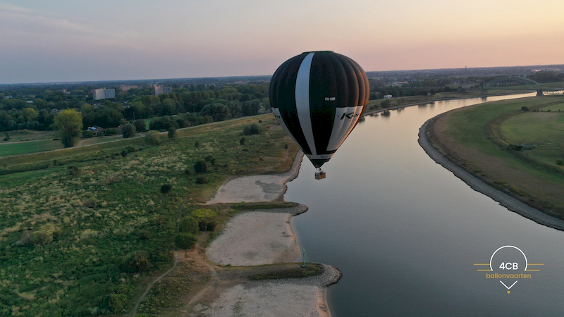 Luchtballon van 4CB Ballonvaarten zwevend laag boven de rivier bij zonsondergang, met uitzicht op oevers en natuur.